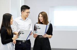 Three people stand indoors, holding and reading brochures, while having a conversation and smiling.