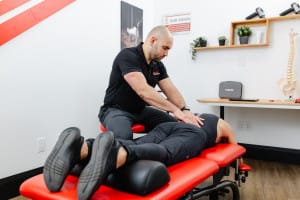 A physical therapist presses on a person's back while they lie face down on a red treatment table in a clinic setting.