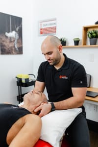 A physiotherapist in a black polo shirt treats a patient's neck as the patient lies on a table in a clinical setting.