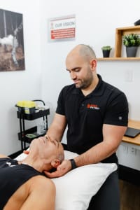 A physical therapist in a black polo shirt treats a patient lying on a table, holding the patient’s head with both hands in a clinical setting.