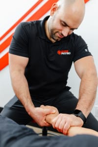 A physical therapist in a black polo shirt performs a manual therapy technique on a patient's forearm in a clinical setting.