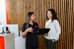 Two women stand and talk at a reception desk; one holds a clipboard, and the other wears a black polo shirt. A wooden slat wall is in the background.