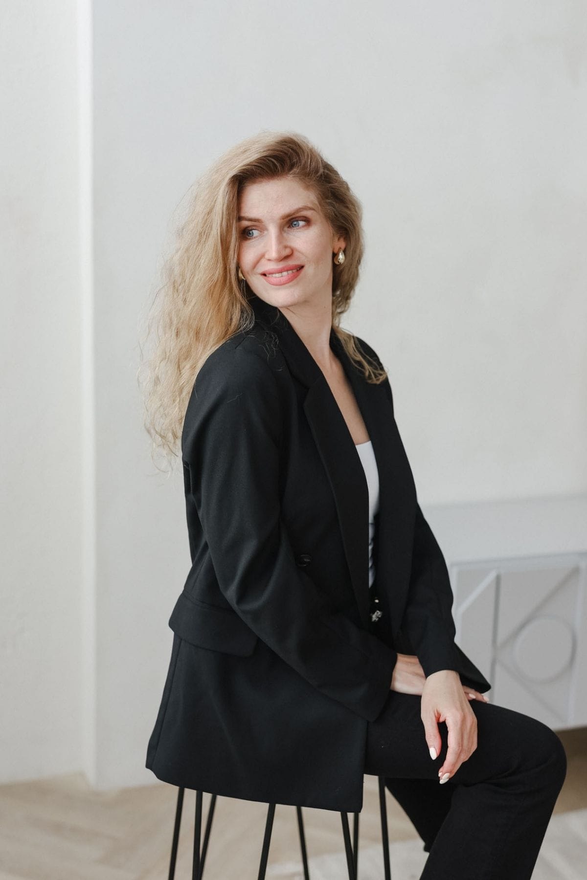 A woman with long blonde hair, part of a creative team, sits on a chair in a minimalist, light-colored room, wearing a black blazer and pants, looking to the side and smiling.