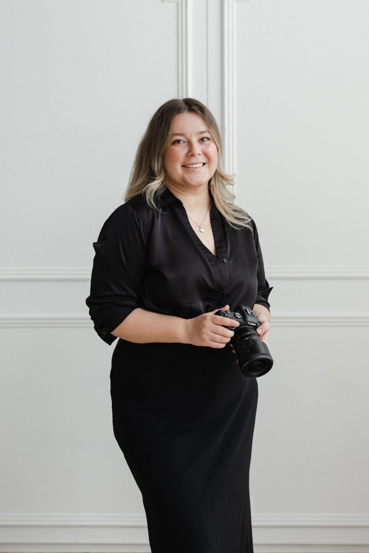 A woman in a black blouse and skirt stands indoors, smiling at the camera with a creative team vibe, holding a camera against a white wall background.