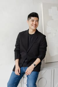 A man from the creative team, wearing a black blazer, dark shirt, and blue jeans, stands indoors, smiling with one hand in his pocket and the other resting on a surface.