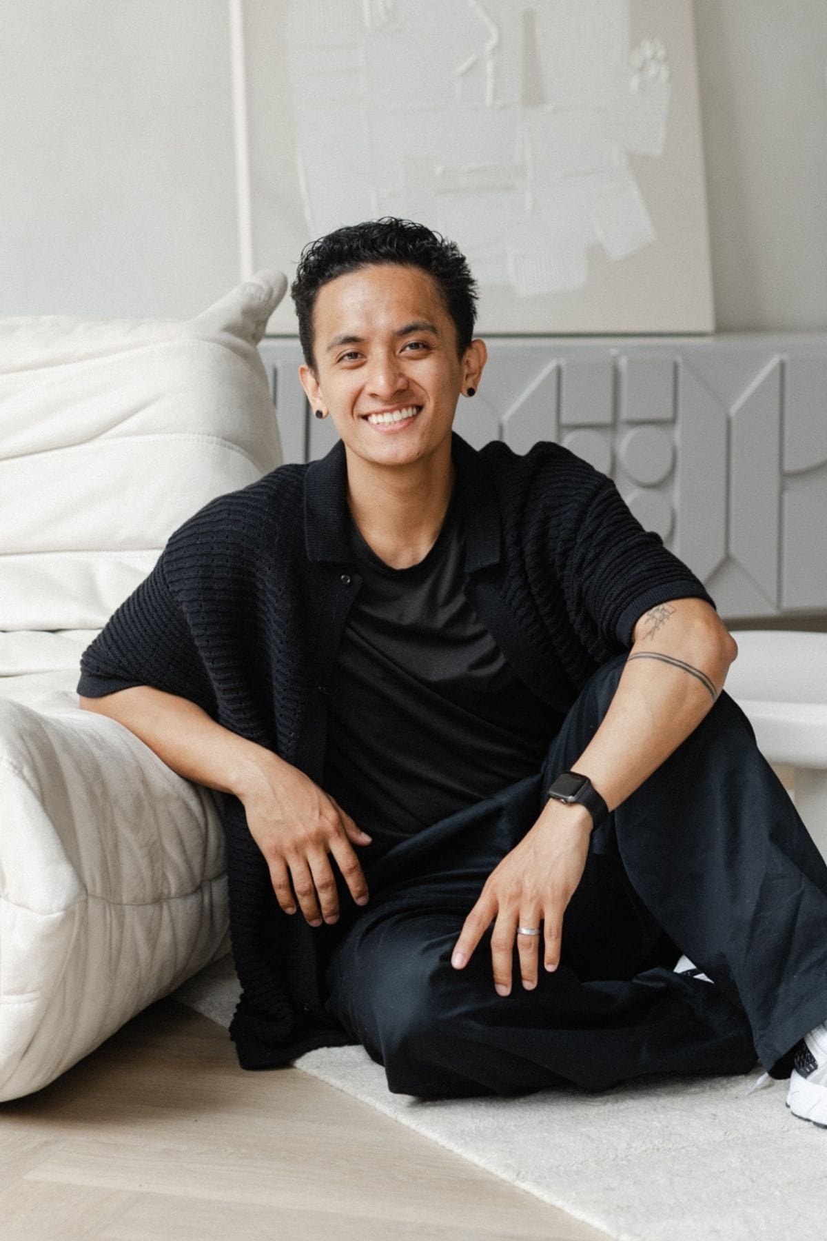 A member of a creative team with short curly hair, wearing a black outfit and a watch, sits on the floor by a white couch, smiling at the camera. Geometric wall art is visible in the background.