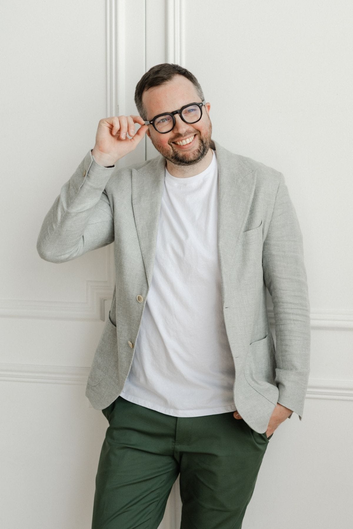 A man with a beard and glasses, part of a creative team, wearing a light gray blazer, white t-shirt, and green pants, stands against a white wall and smiles at the camera.
