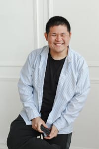 A person with short dark hair in a black t-shirt and striped shirt sits on a stool, smiling and holding a smartphone, ready to connect with their creative team against a plain light-colored background.