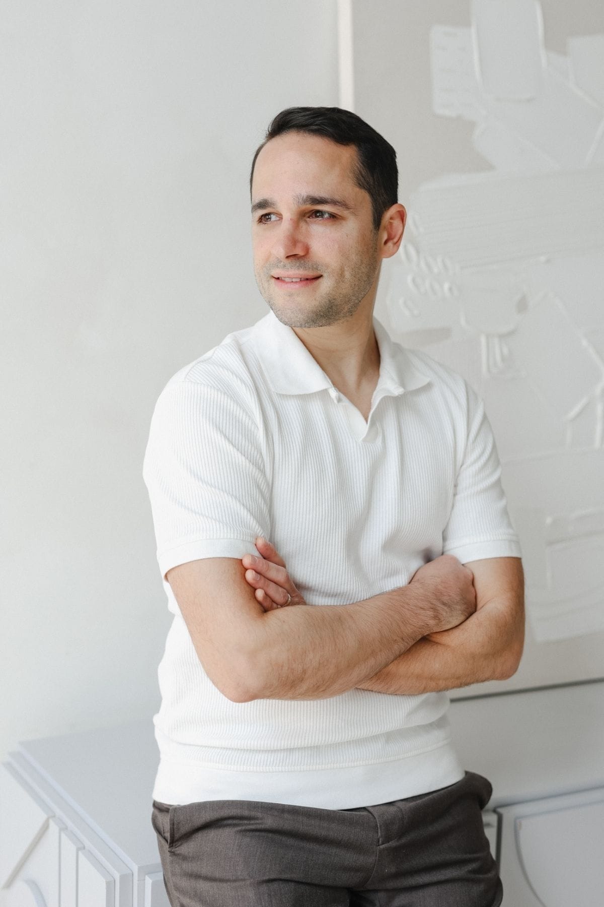 Man with short dark hair wearing a white short-sleeve shirt and dark pants stands with arms crossed, looking to the side in a bright indoor setting, appearing thoughtful—perhaps reflecting on ideas for his creative team.