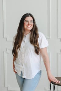 A woman with long brown hair, glasses, and a white shirt stands next to a stool, smiling in front of a white paneled wall—ready to collaborate with the creative team.