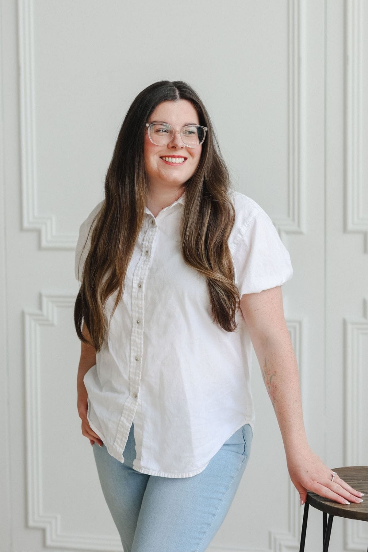 A woman with long brown hair, glasses, and a white shirt stands next to a stool, smiling in front of a white paneled wall—ready to collaborate with the creative team.