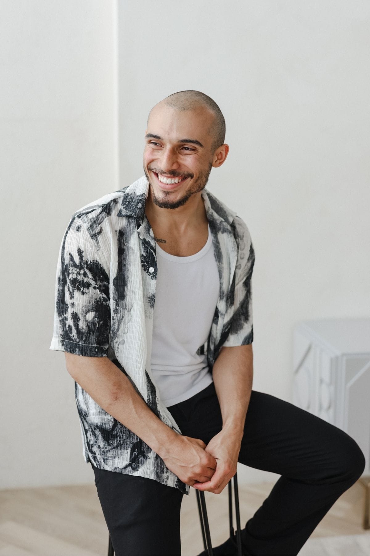 A smiling man with a shaved head, part of a creative team, wears a white tank top and a black-and-white patterned short-sleeve shirt as he sits on a stool against a plain background.