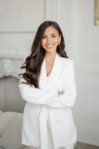 A woman with long brown hair wearing a white suit is standing indoors with her arms crossed, smiling confidently at the camera—a true reflection of a creative team’s dynamic spirit.
