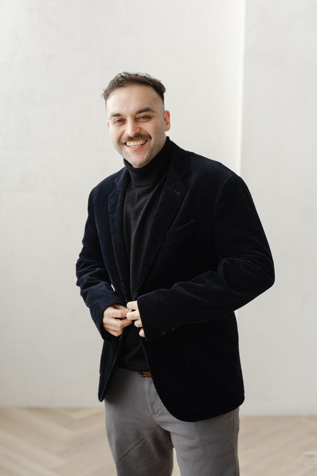 Man with short hair and mustache, part of a creative team, wears a black blazer and turtleneck, standing indoors and smiling while buttoning his jacket.