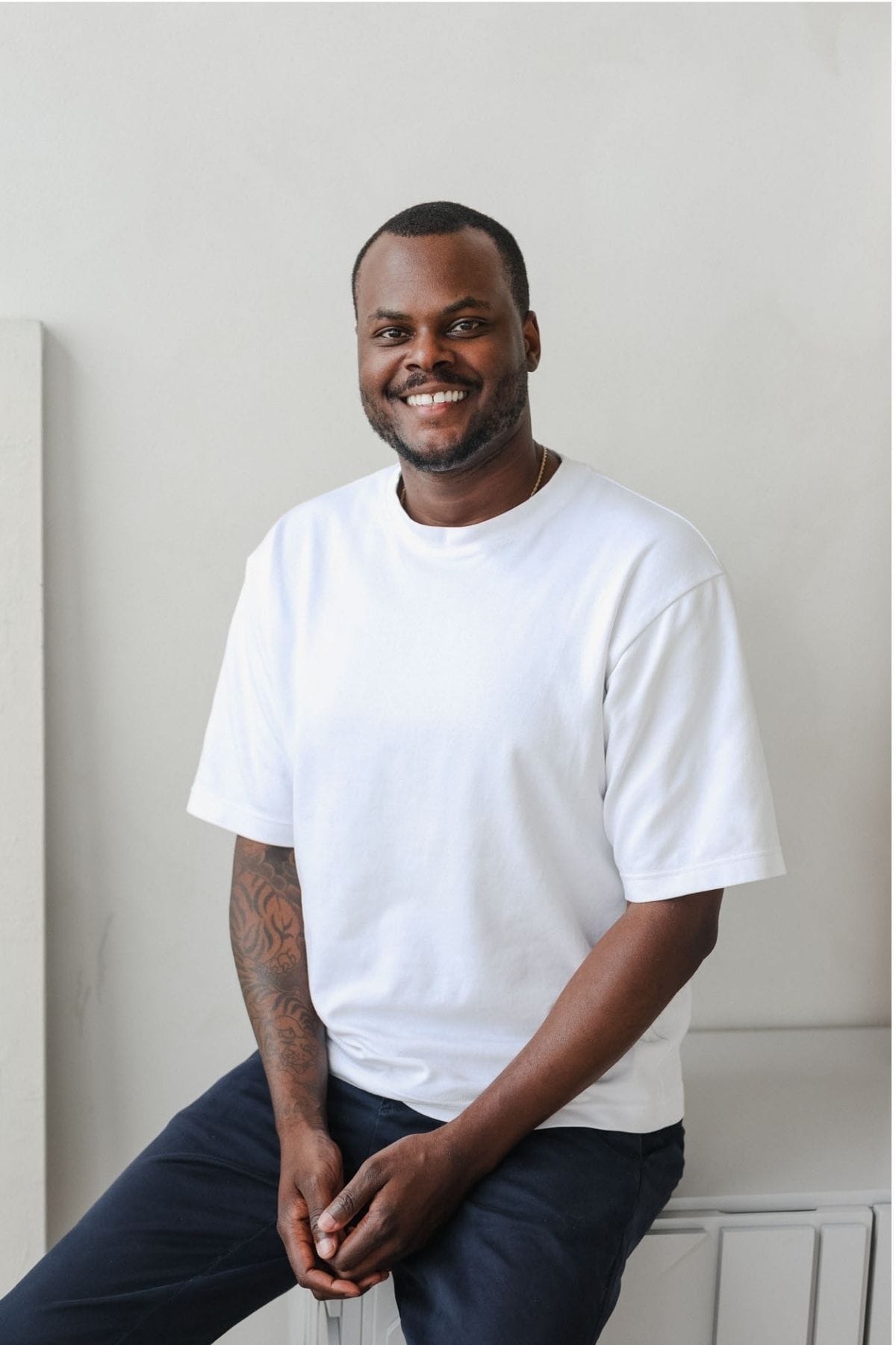 A man with short hair and a beard, wearing a white t-shirt and dark pants, sits on a stool in front of a light gray background, smiling at the camera—ready to join the creative team.