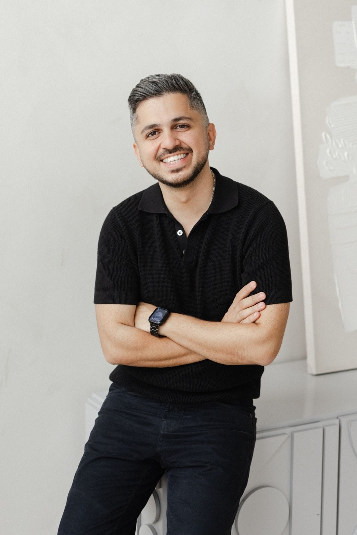 A man with short gray hair and a beard, wearing a black polo shirt and smartwatch, sits on a white cabinet with arms crossed and smiles at the camera, embodying the confidence of a creative team member.