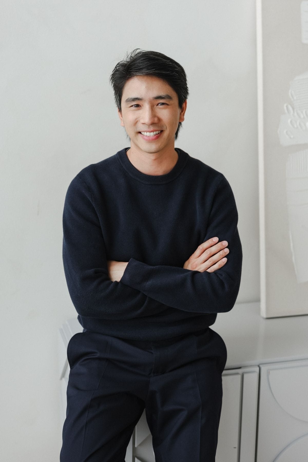 A man with short black hair, wearing a dark sweater and pants, sits on a cabinet with his arms crossed and smiles at the camera, embodying the confident spirit of a creative team member.