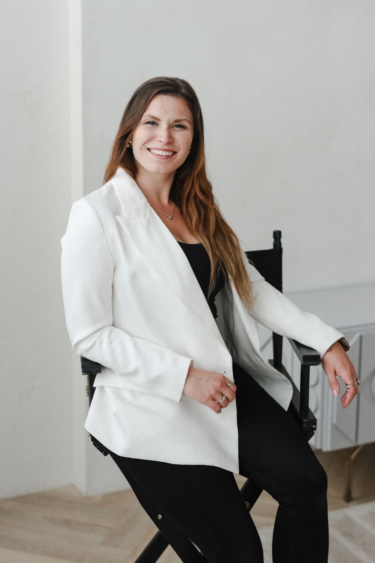 A woman with long brown hair, wearing a white blazer and black pants, sits on a chair and smiles at the camera in a minimal, light-colored room, reflecting the confidence of a creative team member.
