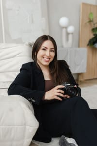 A woman in a black suit sits on a light-colored sofa, smiling and holding a Canon camera in her hands—ready to capture moments for her creative team.