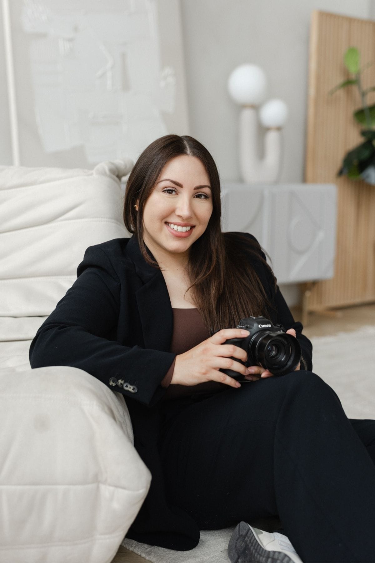A woman in a black suit sits on a light-colored sofa, smiling and holding a Canon camera in her hands—ready to capture moments for her creative team.