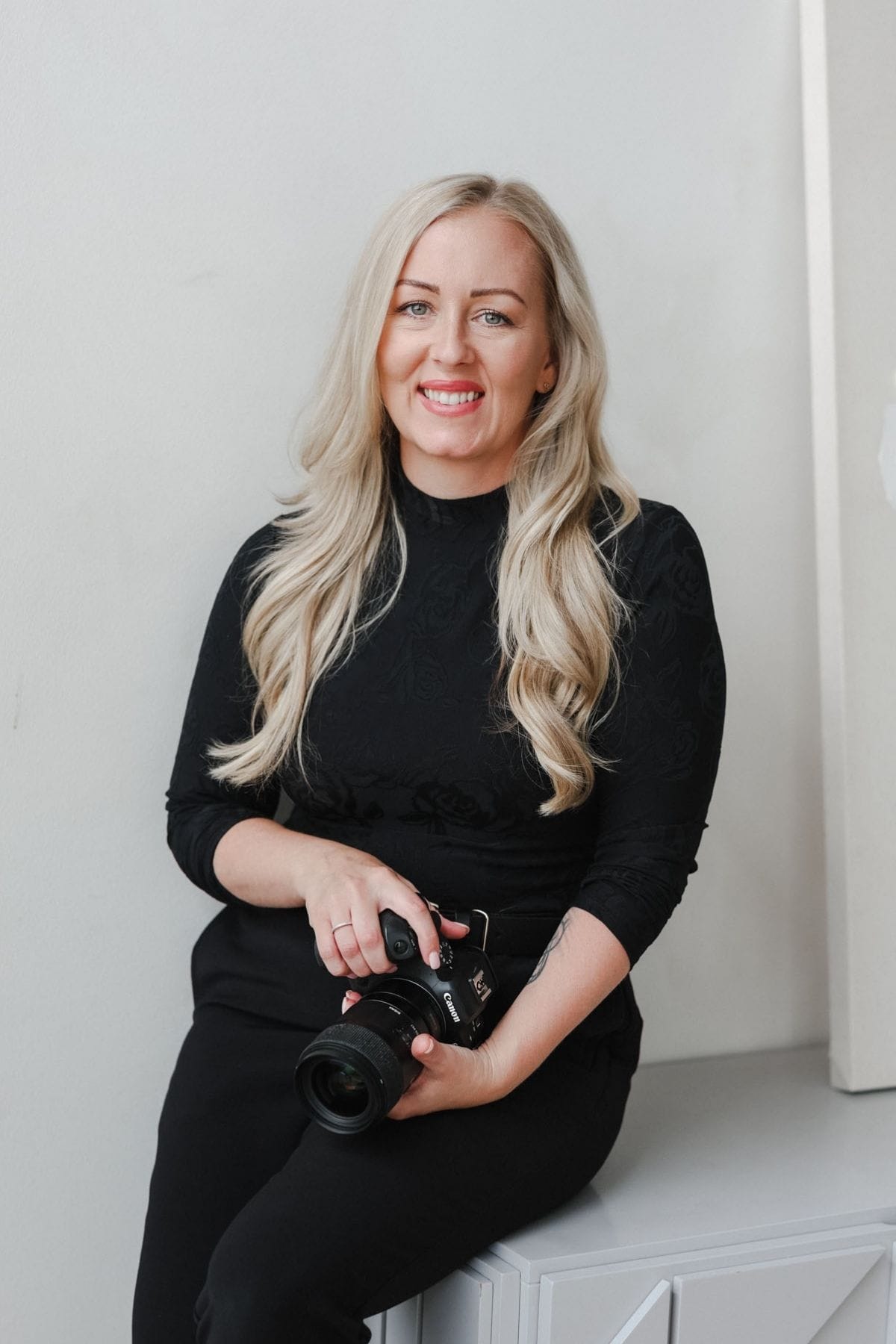 A woman with long blonde hair, part of a creative team, wears a black outfit as she sits on a light-colored surface, holding a camera and smiling at the camera.