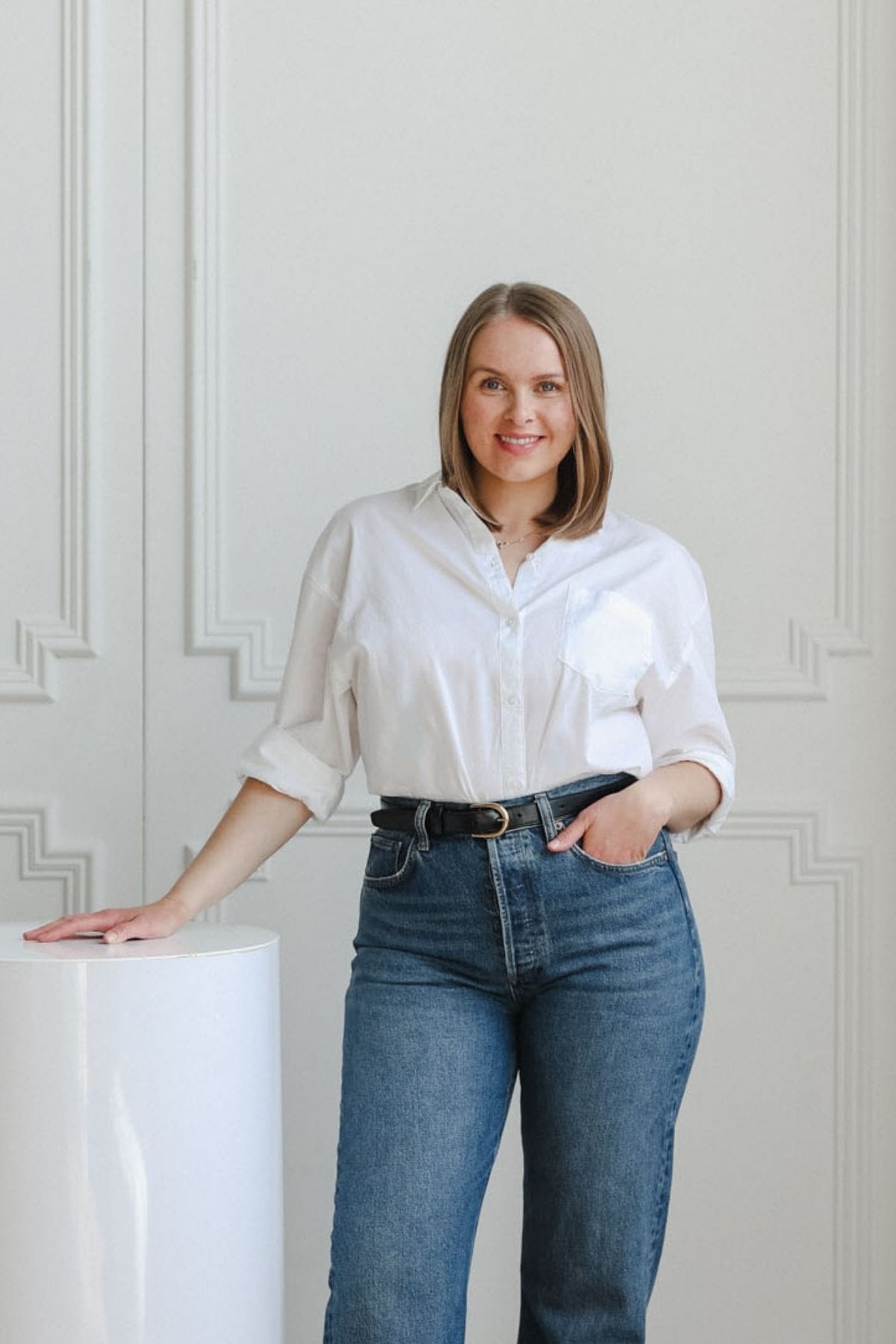 A woman in a white shirt and blue jeans stands indoors, resting one hand on a white cylindrical pedestal and the other in her pocket, against a white paneled wall—poised as if ready to join a creative team.