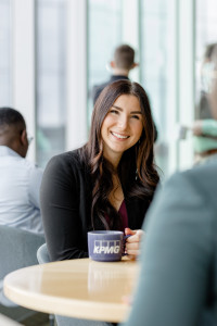 A woman with long brown hair sits at a table holding a KPMG mug, smiling at someone across from her in a modern office setting, discussing high-impact content strategies for marketing on a budget.