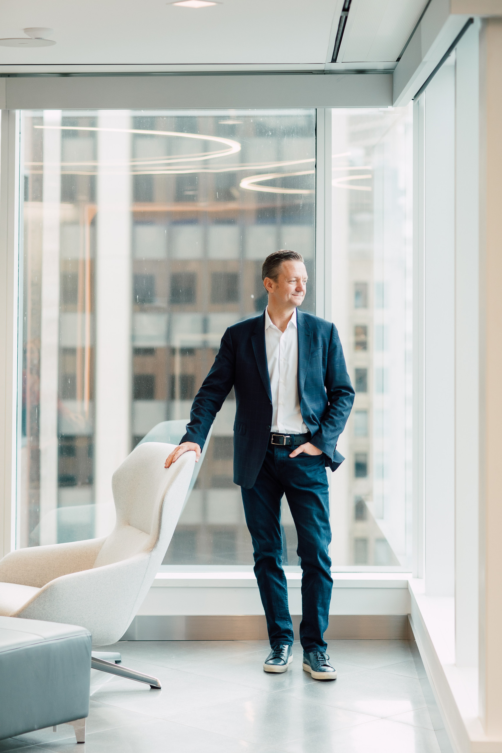 A man in a navy blazer and jeans stands by a large window in a modern office, perhaps reflecting on marketing on a budget as he rests one hand on a white chair and gazes outside.