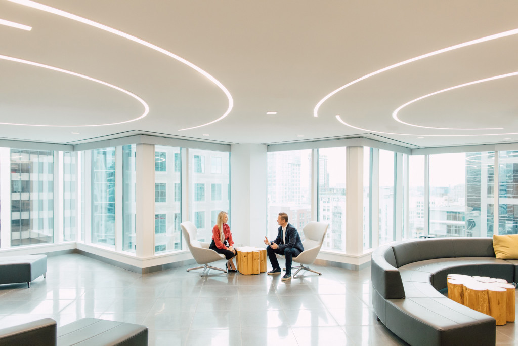 Two people sit and talk in a modern, bright office lounge with large windows, city views, and contemporary furniture, brainstorming high-impact content strategies for marketing on a budget.
