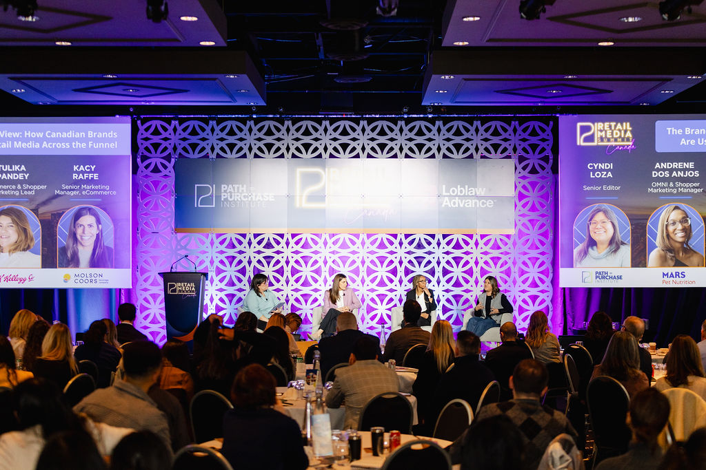 A panel of four speakers discusses retail media on a stage at a conference, with an audience seated and large screens displaying speakers' names and titles.