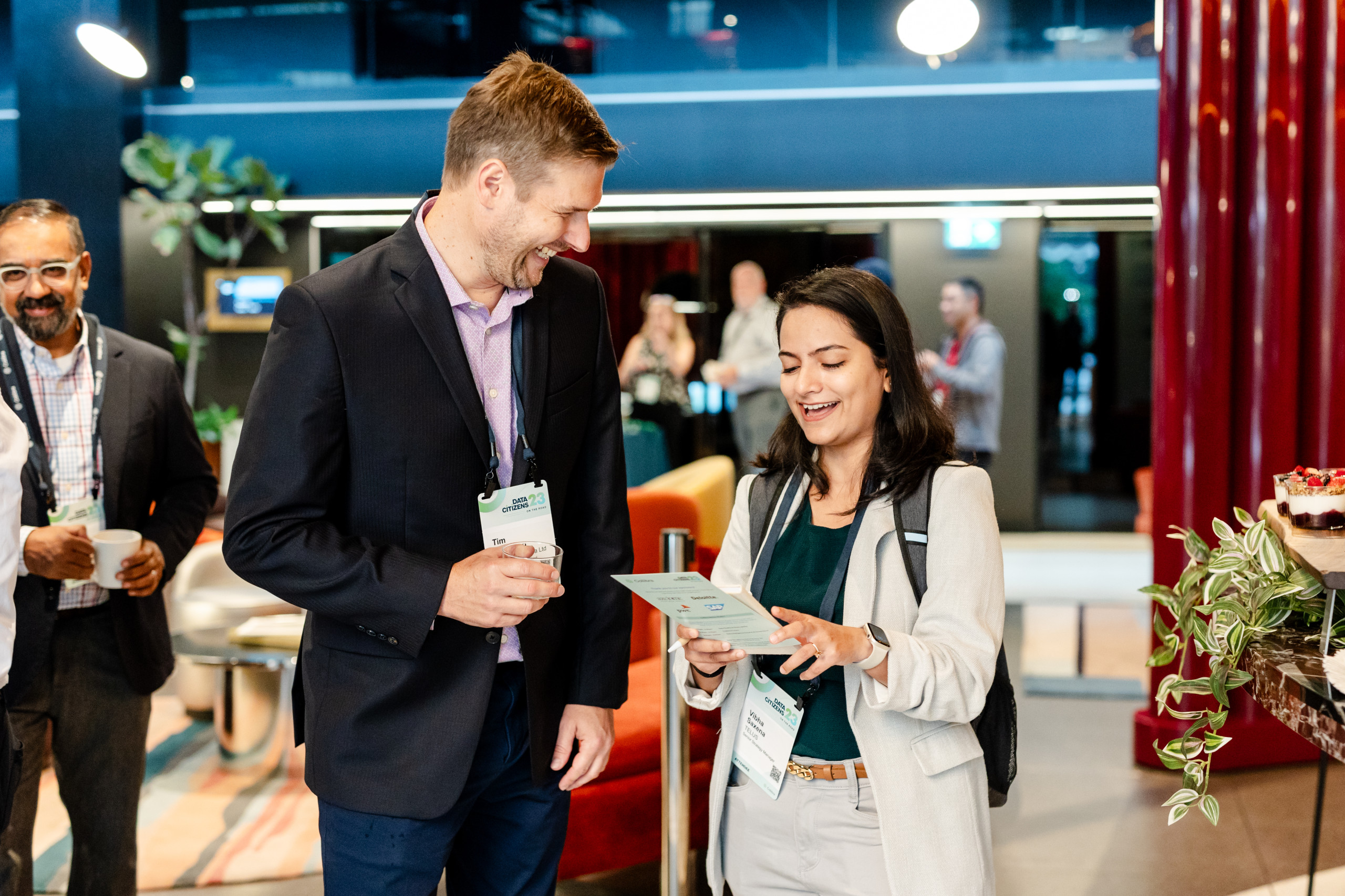 A man and a woman stand indoors at a networking event, smiling and looking at a pamphlet, as others mingle in the modern decor—capturing the spirit of events 2026 and the latest photography trends.