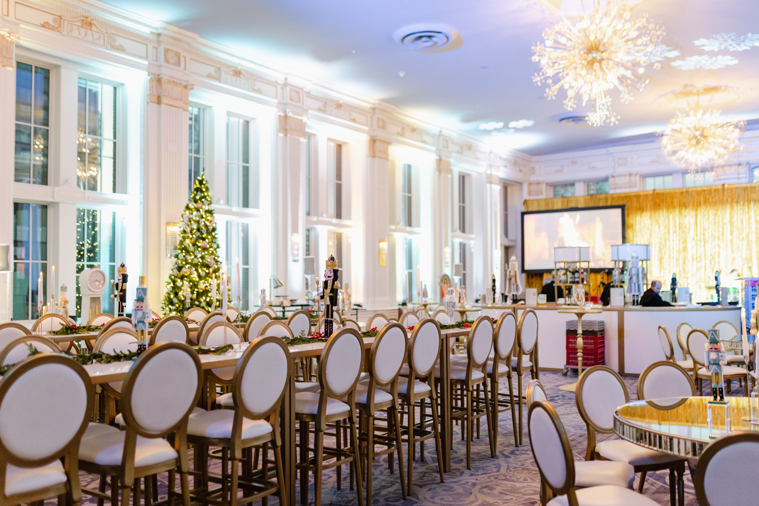 A decorated event hall with round-backed chairs, round tables, a Christmas tree, chandeliers, and a bar area in the background.