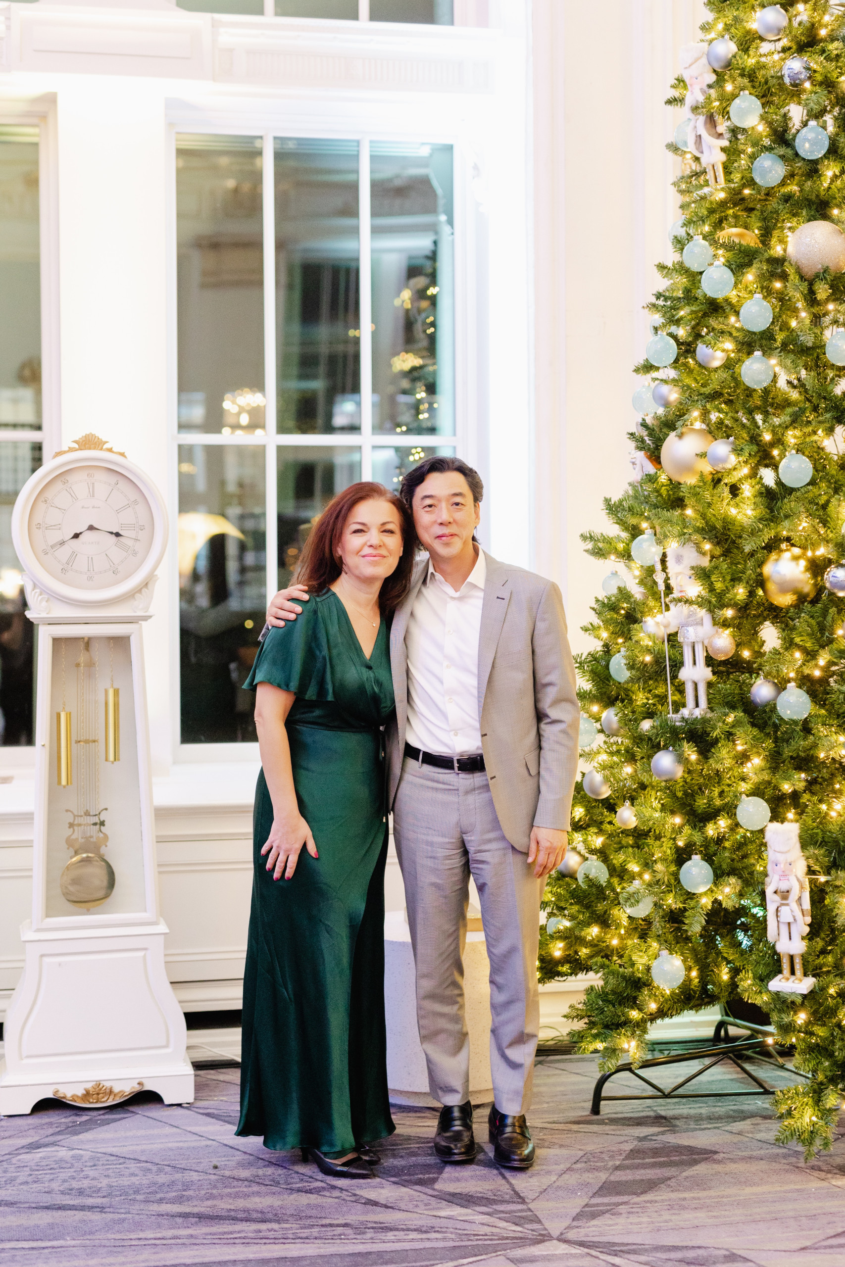 A woman in a green dress and a man in a gray suit stand together by a decorated Christmas tree and a white grandfather clock in a brightly lit room.