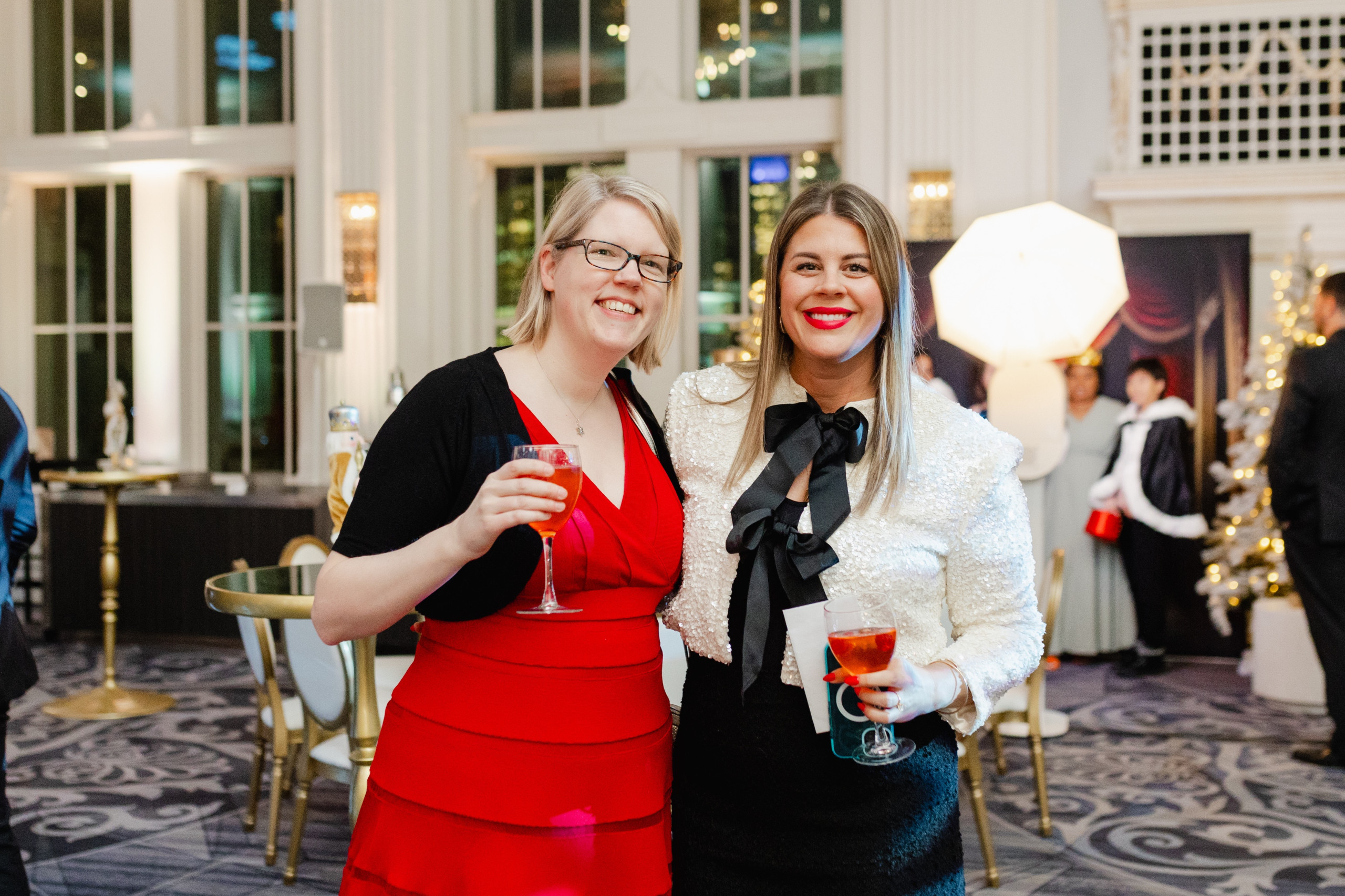 Two women in formal attire smile and pose with drinks at an indoor event with festive decor and people in the background.