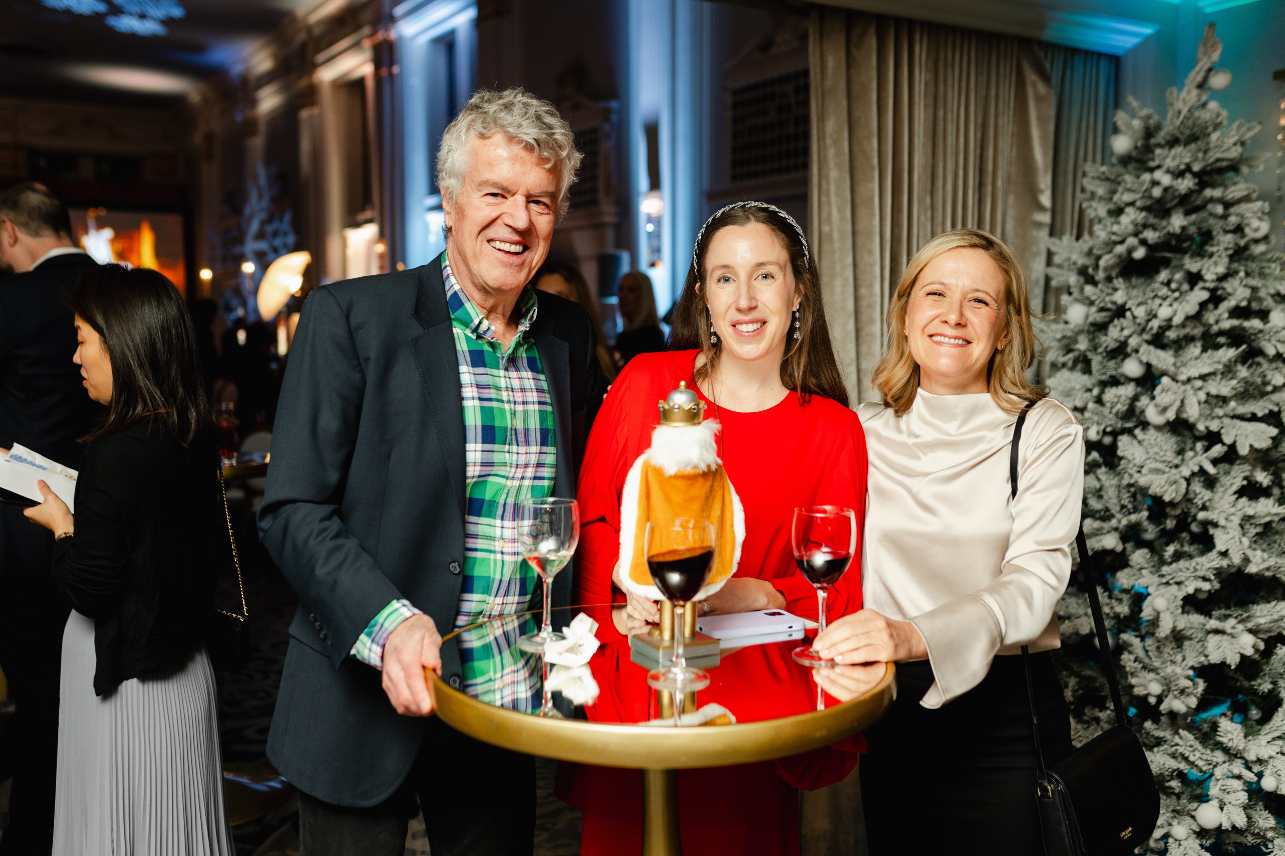 Three adults stand around a round table with wine glasses at an indoor holiday party, with a decorated tree and festive decor in the background.