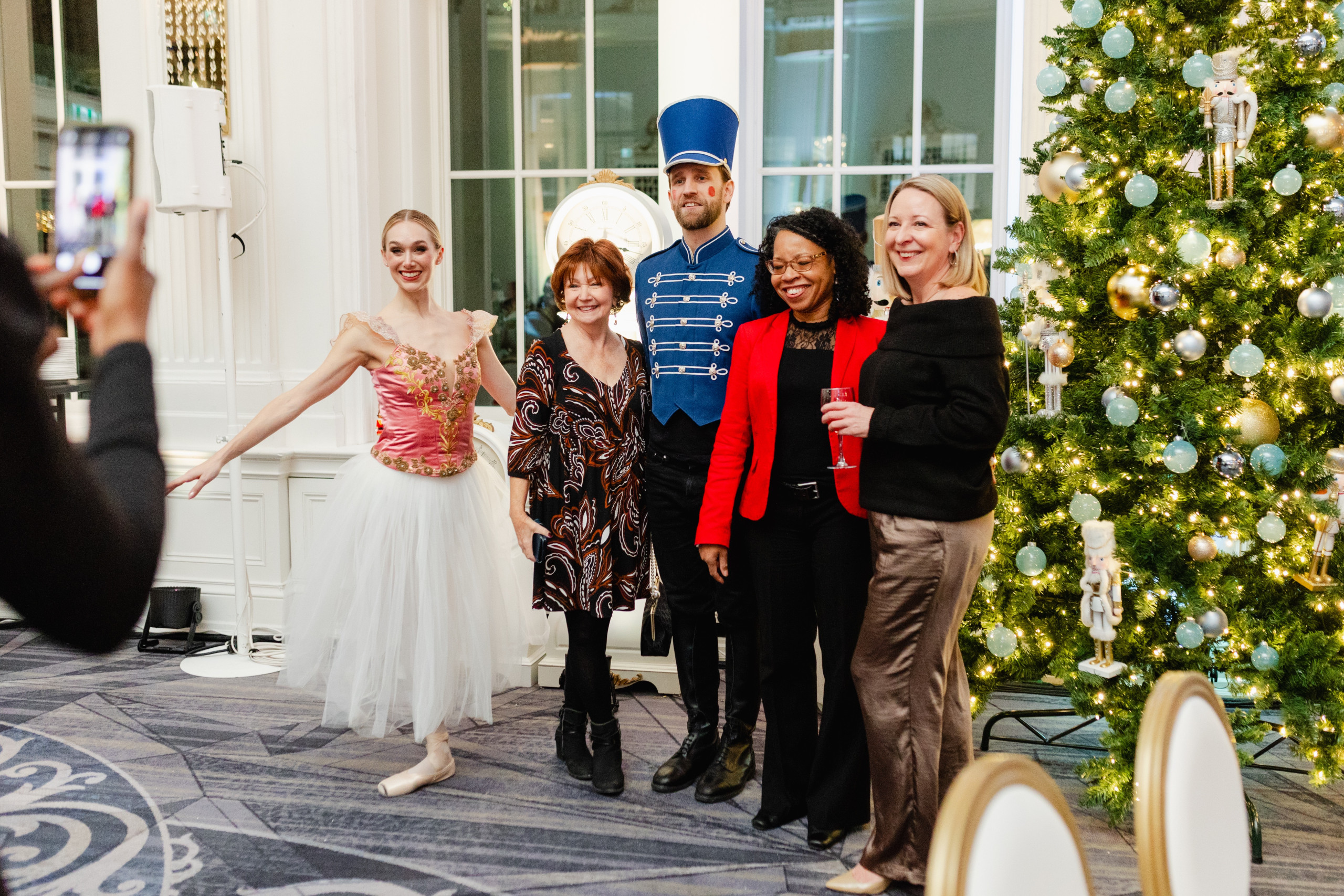 Five people pose for a photo at a festive event; one is dressed as a ballerina, another as a soldier, standing beside a decorated Christmas tree indoors.