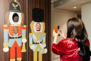 A woman takes a photo of another woman posing with her face through a cutout in a wooden nutcracker soldier standee.