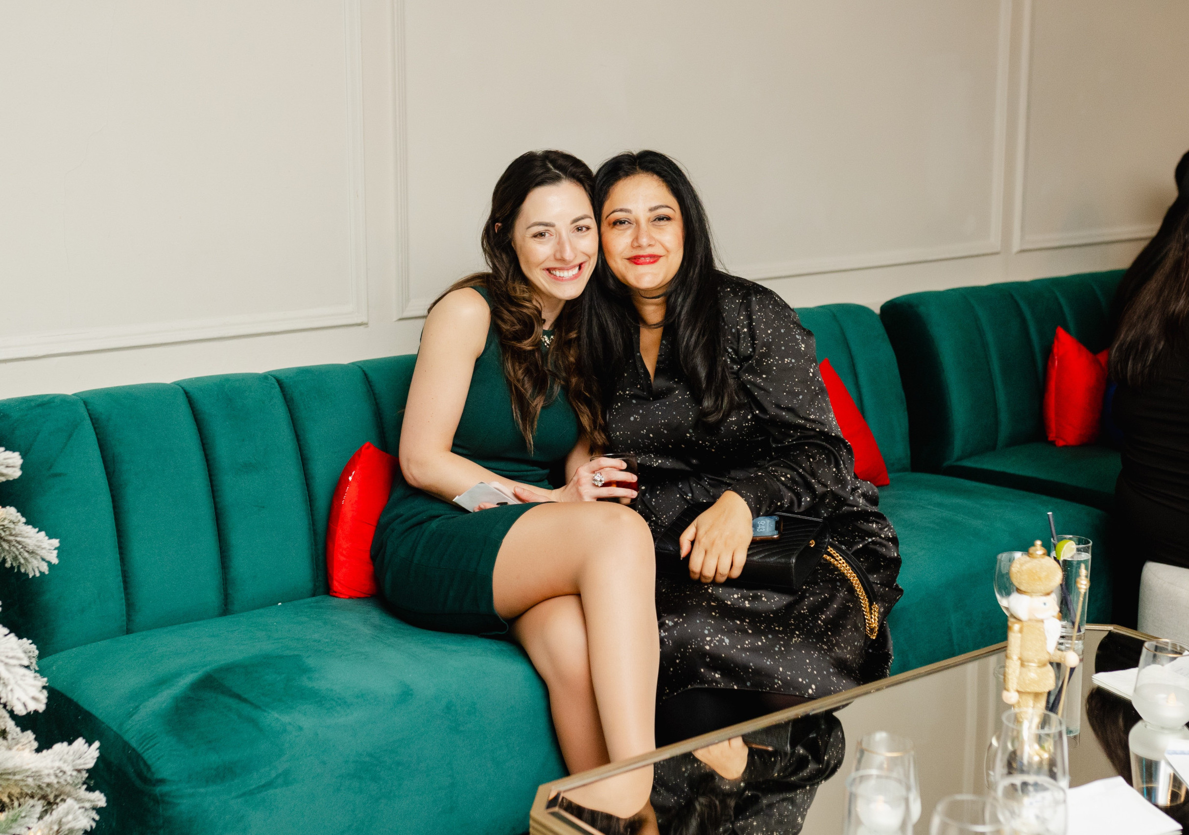 Two women sitting close together on a green couch, smiling at the camera, with red cushions and a decorated tree partially visible on the left.