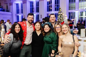 A group of seven adults, some dressed festively, pose and smile together at a holiday party in a decorated indoor venue with a lit Christmas tree in the background.