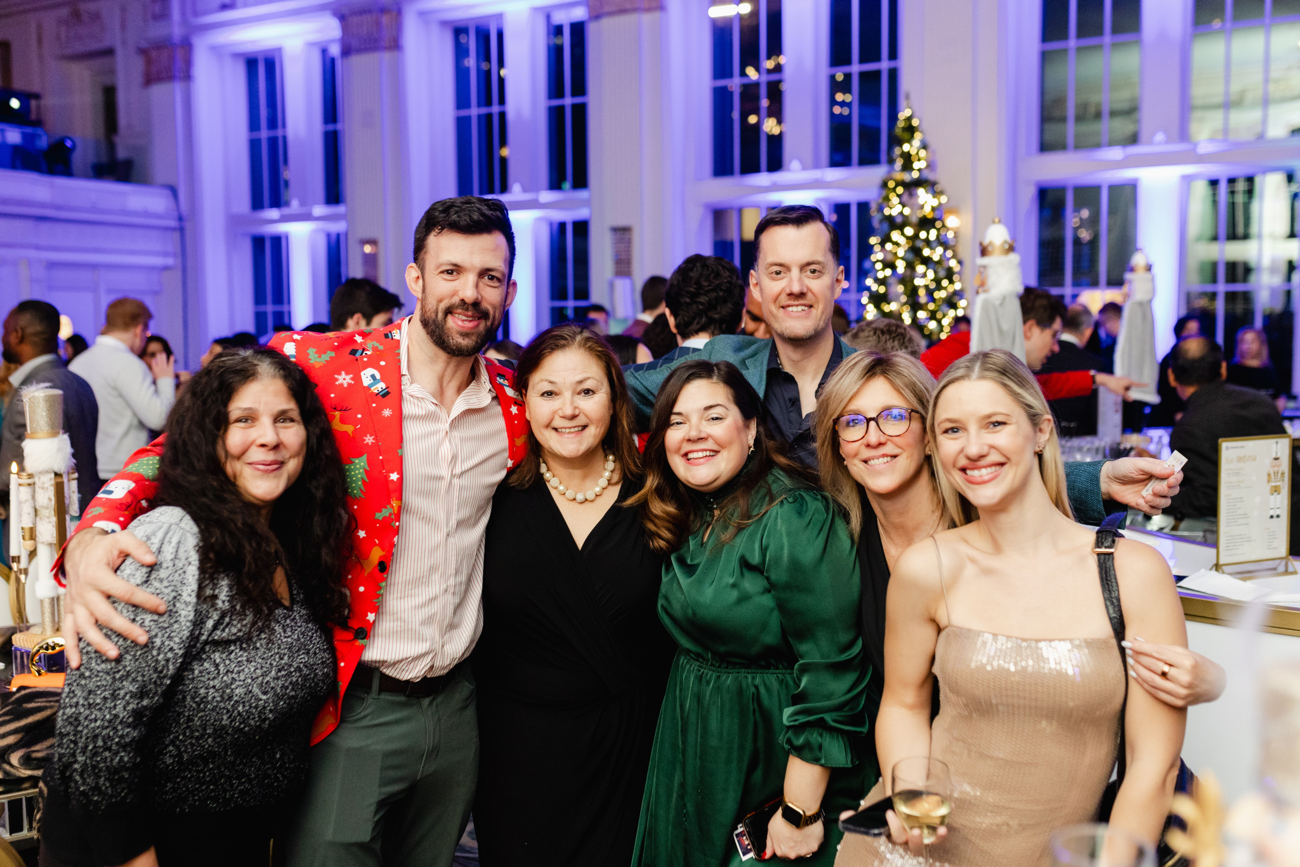 A group of seven adults, some dressed festively, pose and smile together at a holiday party in a decorated indoor venue with a lit Christmas tree in the background.