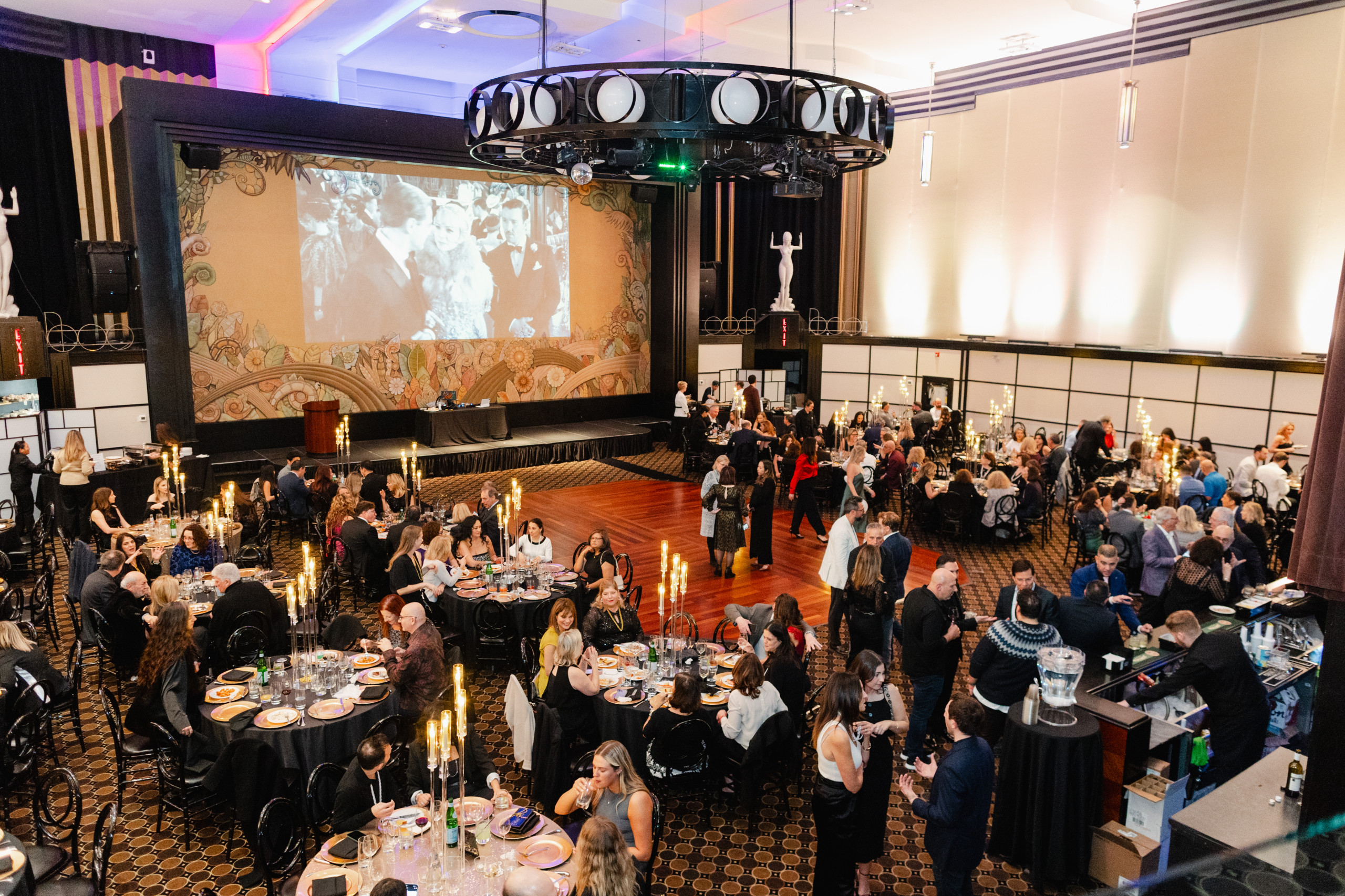 A large group of people dine and socialize at round tables in a decorated banquet hall with a stage, large screen, and candlelit centerpieces.