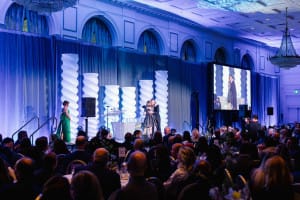 A formal event in a grand ballroom shows a presenter on stage addressing an audience, with large decorative columns and a projection screen displaying the stage.