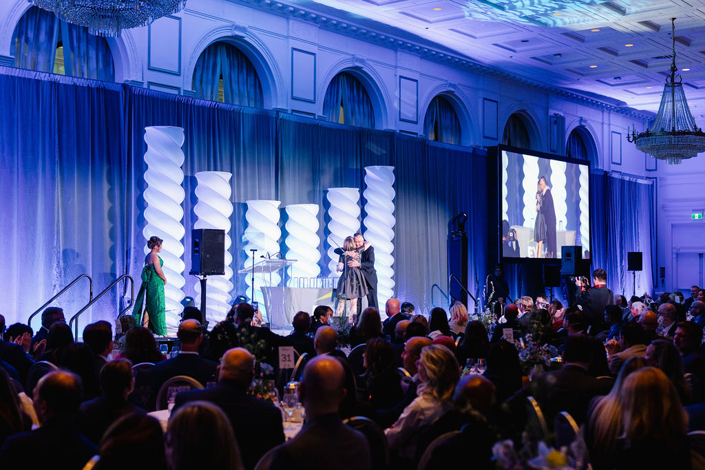A formal event in a grand ballroom shows a presenter on stage addressing an audience, with large decorative columns and a projection screen displaying the stage.