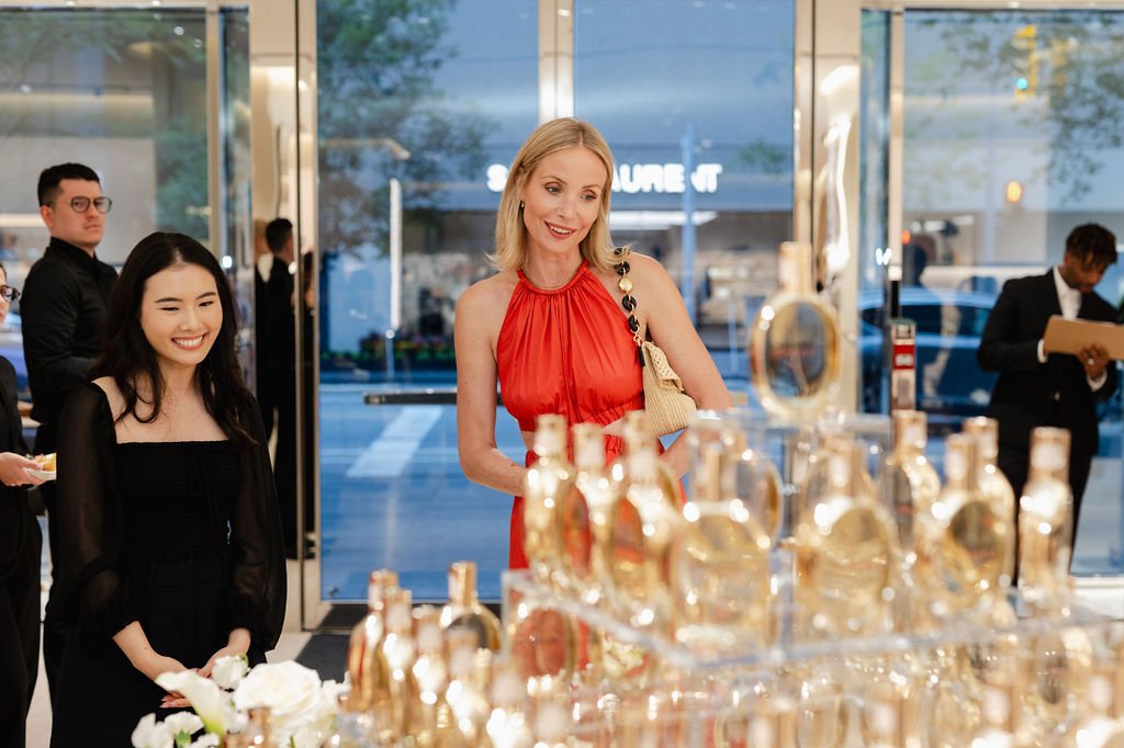 Two women stand in front of a display of gold perfume bottles inside a well-lit store, with other people visible in the background.