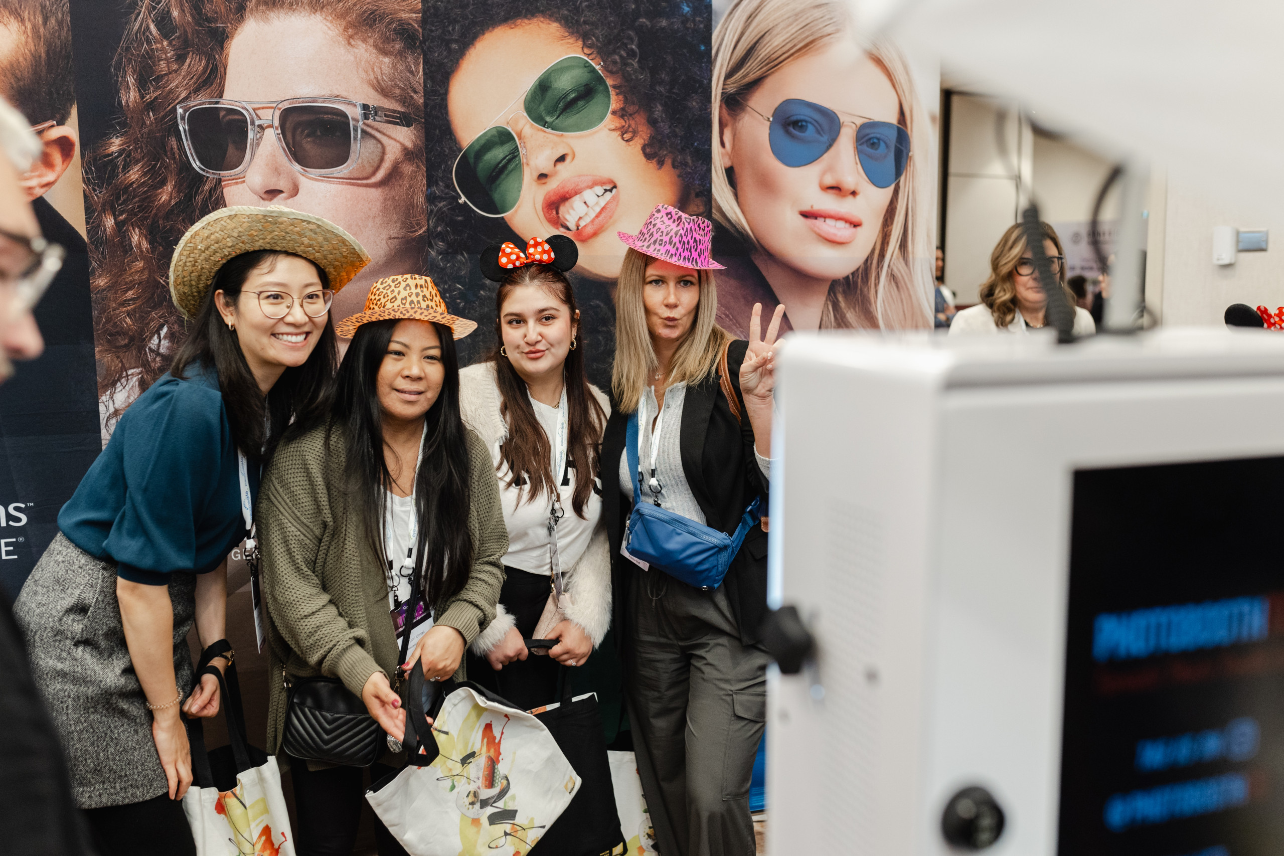 Four women wearing colorful hats and posing for a photo booth in front of a sunglasses advertisement backdrop at an indoor event.