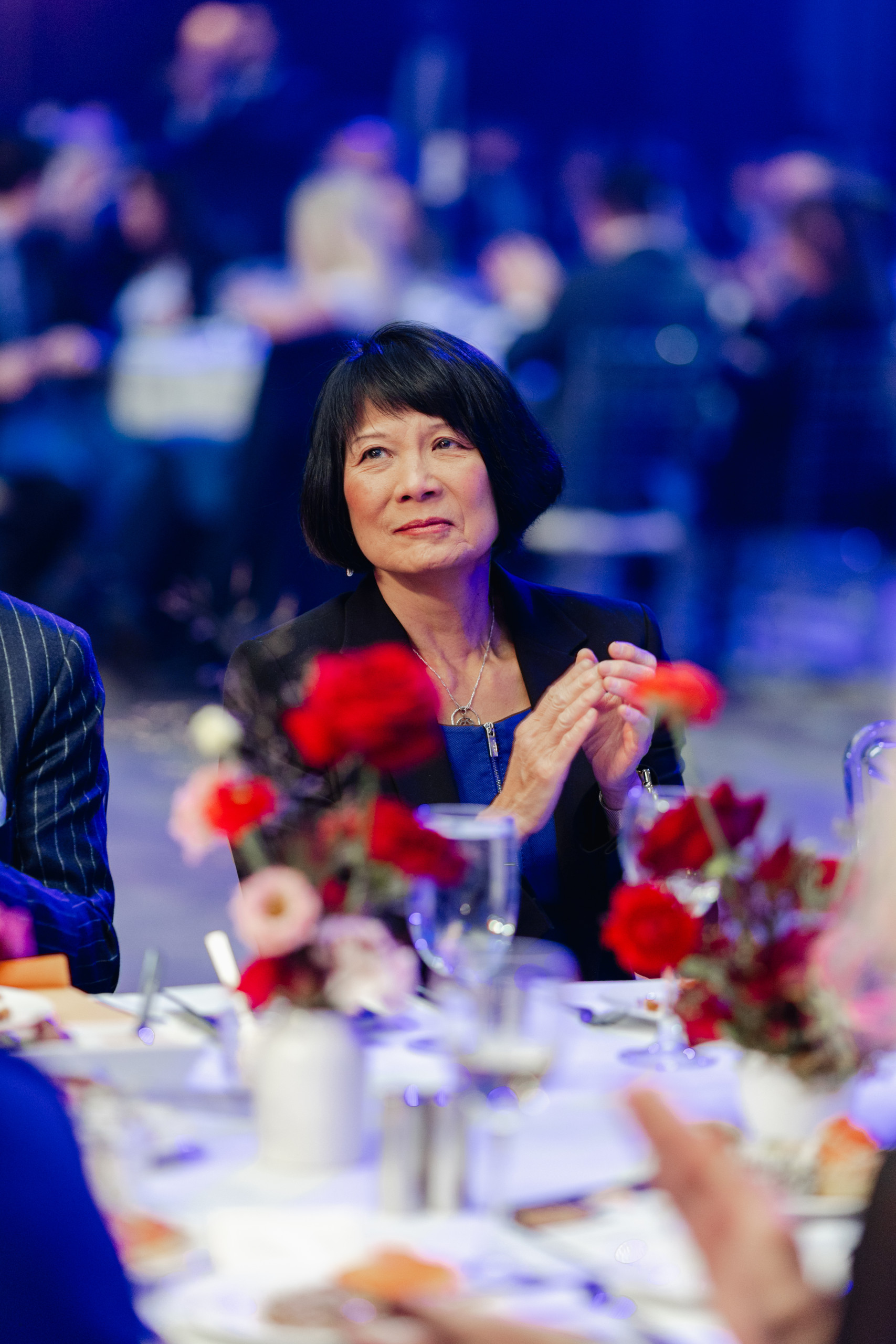 A woman with short black hair sits at a round table with floral centerpieces, clapping her hands at a formal event with other people in the background.