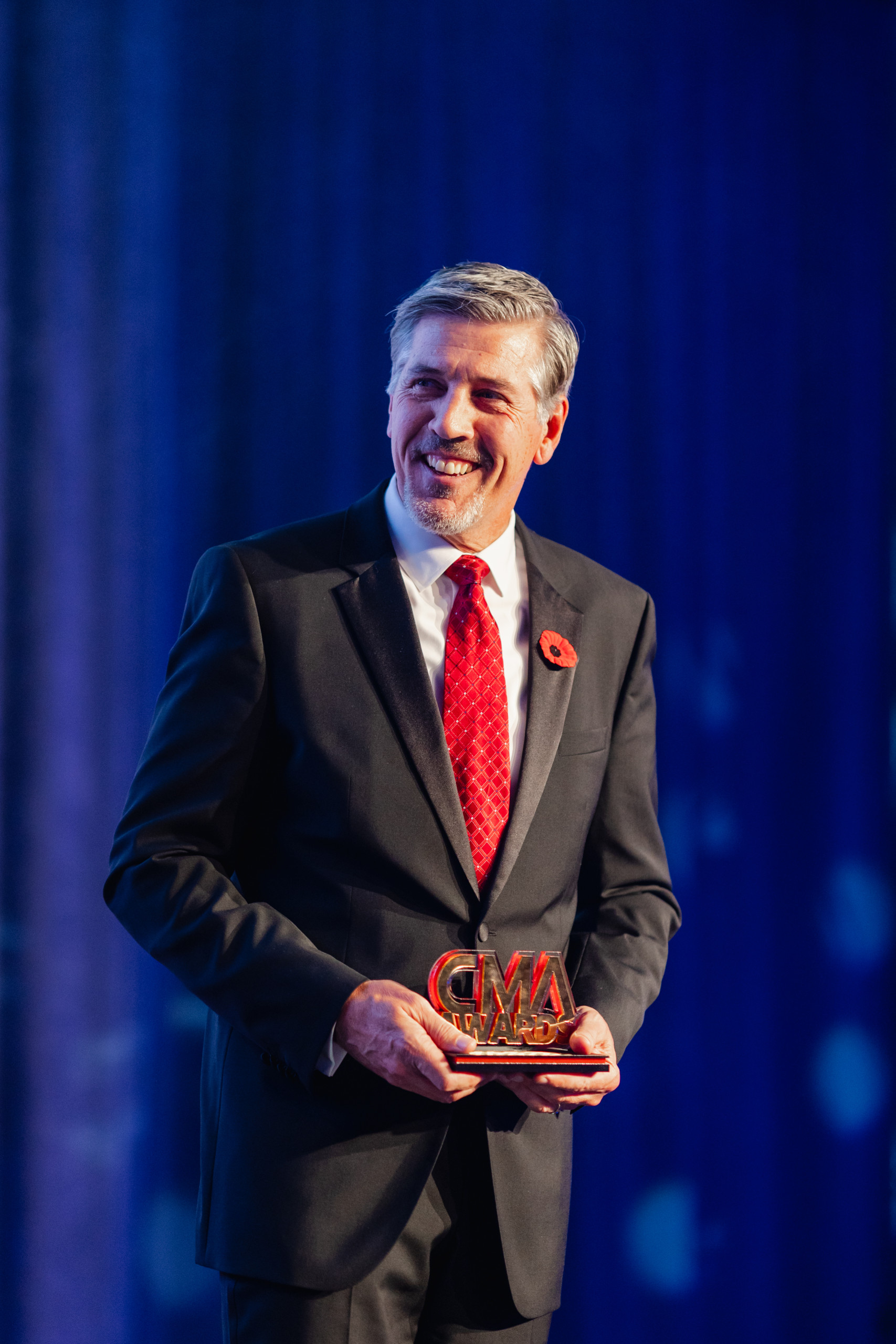 A man in a suit and red tie holds a CMA Awards trophy while standing in front of a blue background.