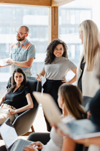 Five people in business casual attire gather in a bright office space, engaged in discussion about marketing on a budget, with some using laptops and notebooks.