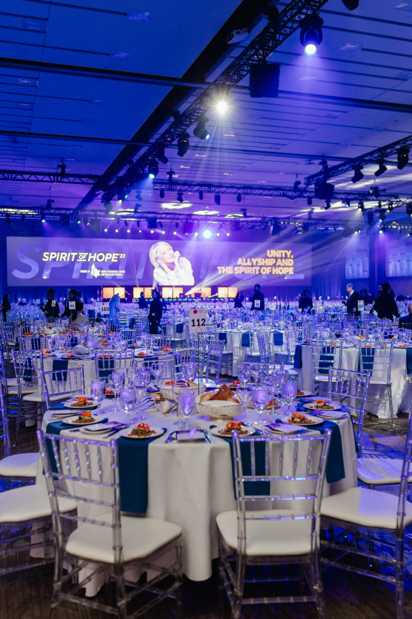 A banquet hall with round tables set for an event, featuring clear chairs, plates of food, and a stage with a large screen reading "SPIRIT of HOPE" and "UNITY, ALLYSHIP AND THE SPIRIT OF HOPE".