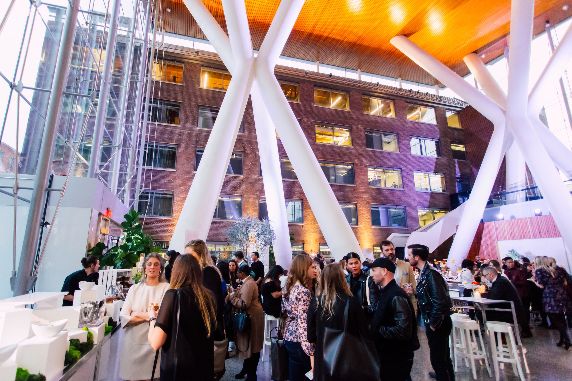 A crowd of people socialize in a modern indoor venue with tall white columns, a glass wall, and exposed brick backdrop.